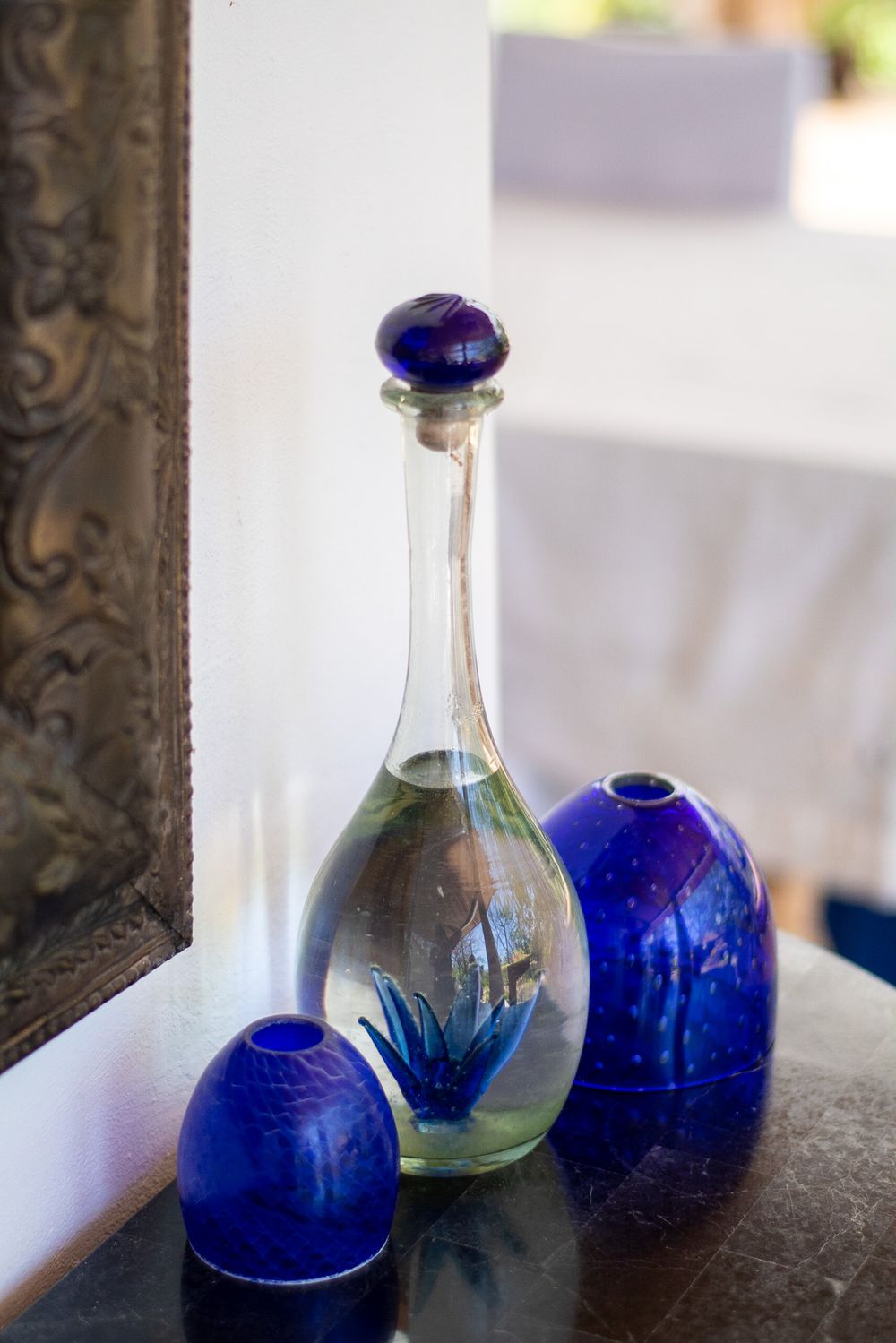Elegant cobalt blue glass vessels and a clear bottle arranged on a dark surface near an ornate frame.