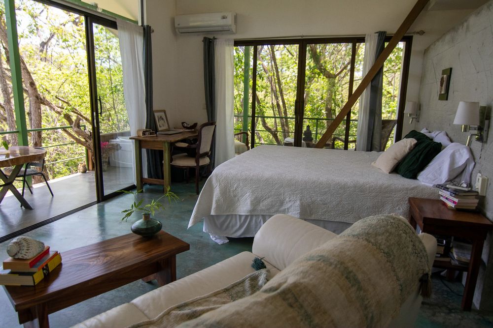 An airy bedroom with large windows overlooking trees, featuring white bedding and wooden furniture.