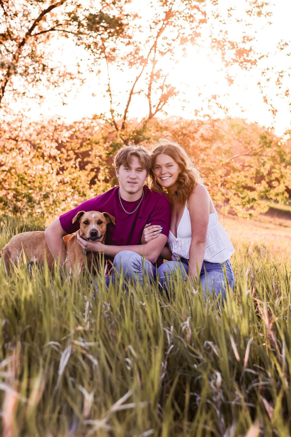A couple poses with their dog in tall grass during golden hour.