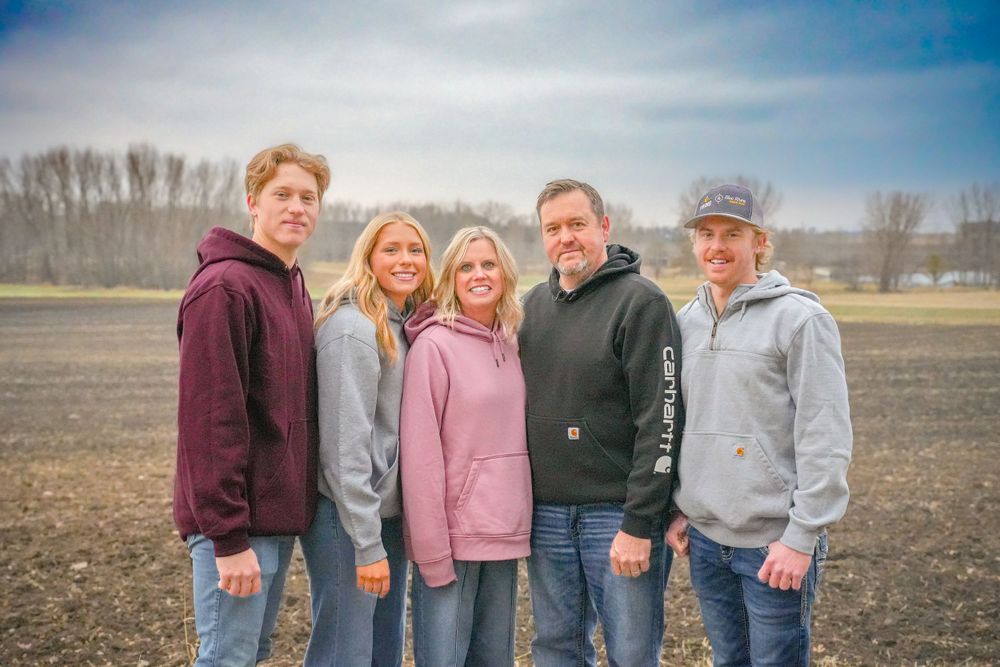 Family of five poses together in casual wear against rural landscape.
