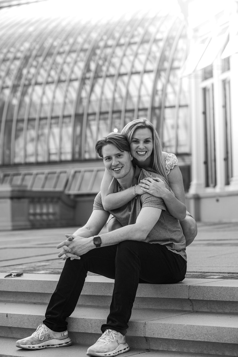 A romantic black and white portrait of a couple embracing on outdoor steps near a modern glass building.