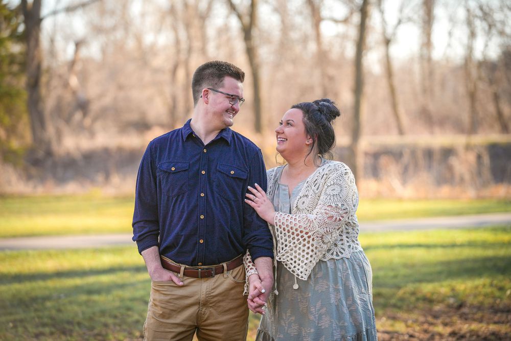Couple smiling and holding hands outdoors in a park during golden hour, wearing casual elegant attire.