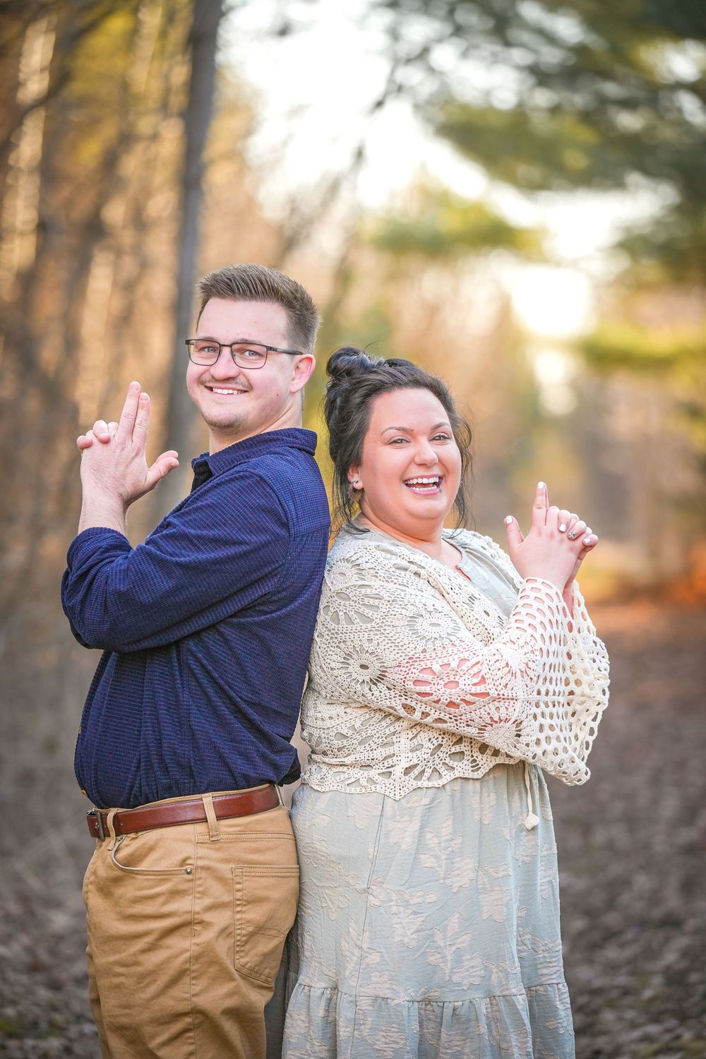 Couple standing back-to-back laughing in autumn forest, man in navy shirt and khakis, woman in lace dress.