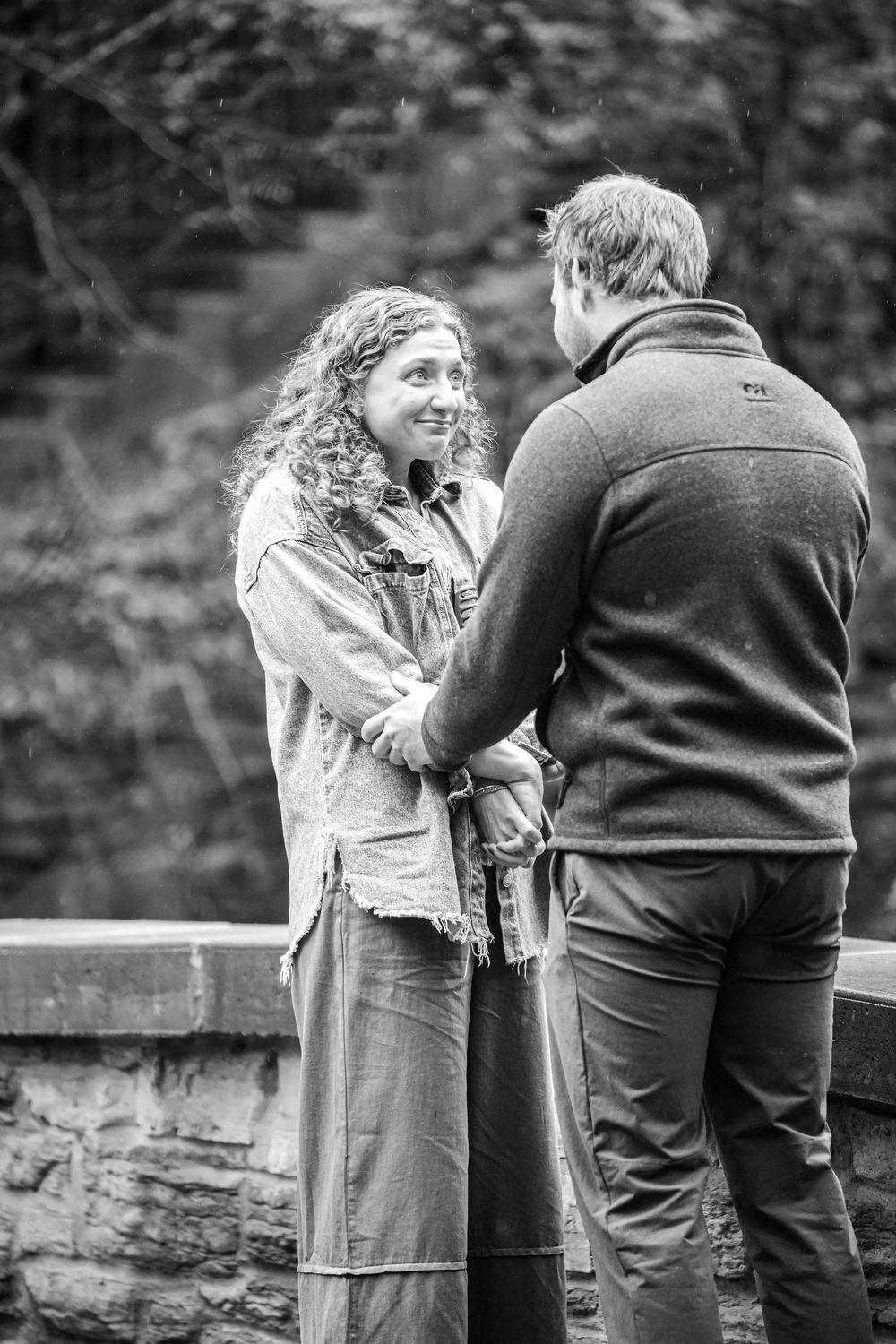Black and white photo of a smiling curly-haired woman holding hands with a man outdoors near a stone wall.