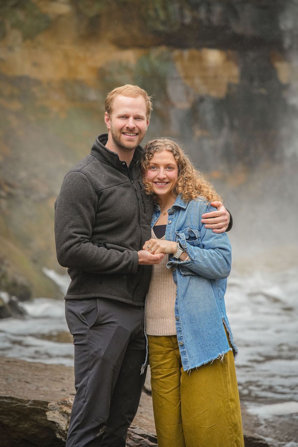 A smiling couple embraces in front of a misty waterfall, the man in gray fleece and woman in denim jacket and yellow pants.