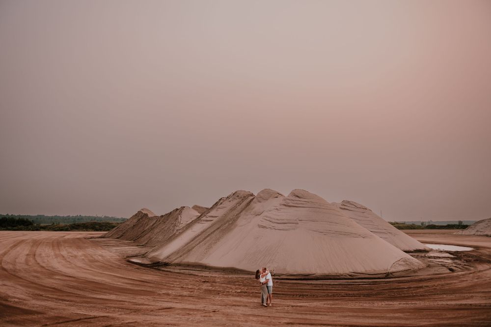 A small figure stands before massive sand dunes under a hazy pink sky at a mining or industrial site.