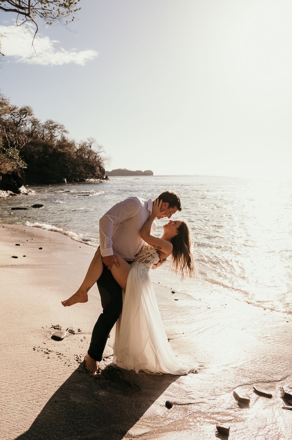 A romantic couple embraces on a sandy beach at sunset with waves lapping at the shore and trees in the background.