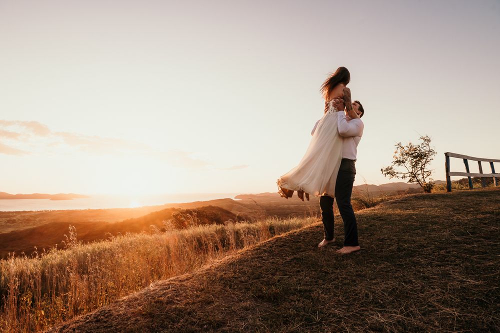 A couple embraces on a hilltop silhouetted against a golden sunset sky with tall grass in the foreground.