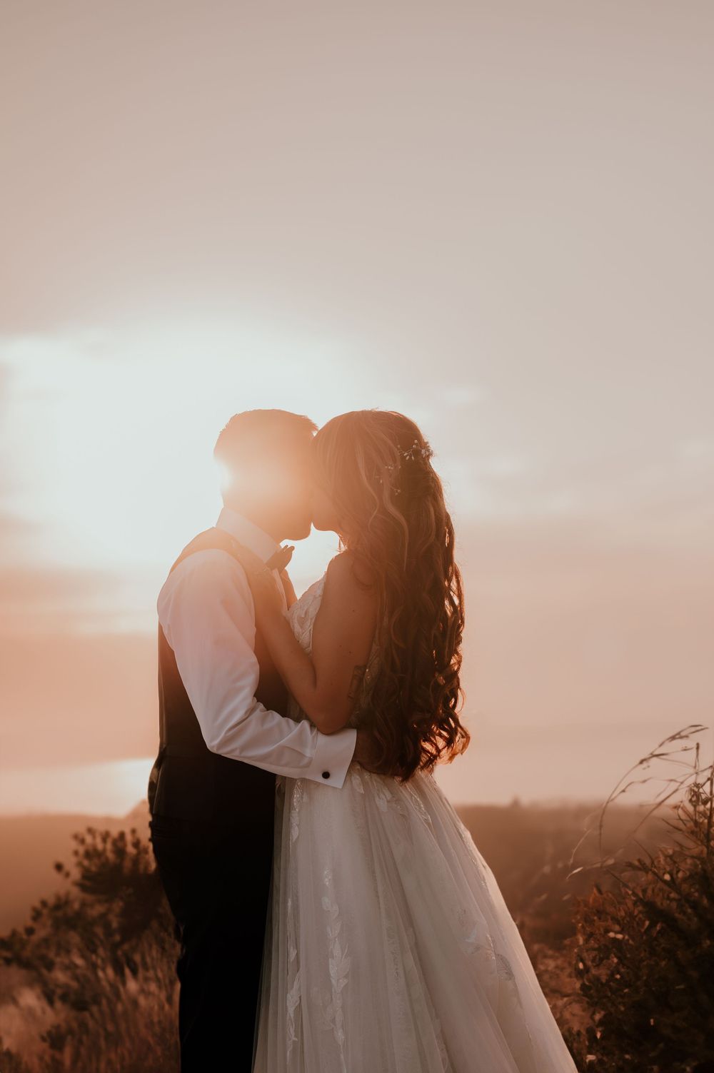 A silhouetted couple in wedding attire share an intimate moment at sunset against a golden sky and mountain landscape. mariés au golden hour