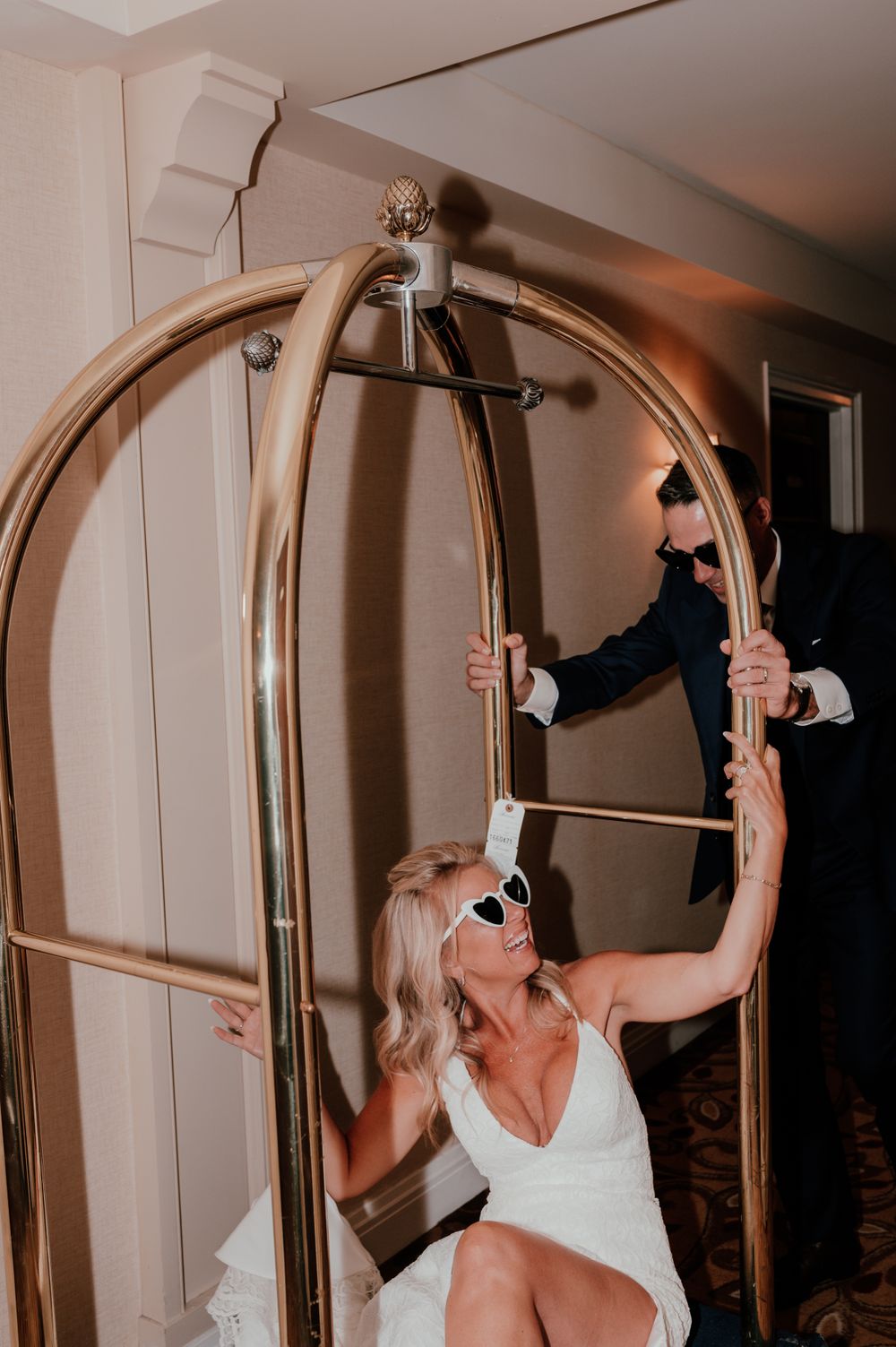 Woman in white dress swinging on a brass luggage cart at a hotel during an event.