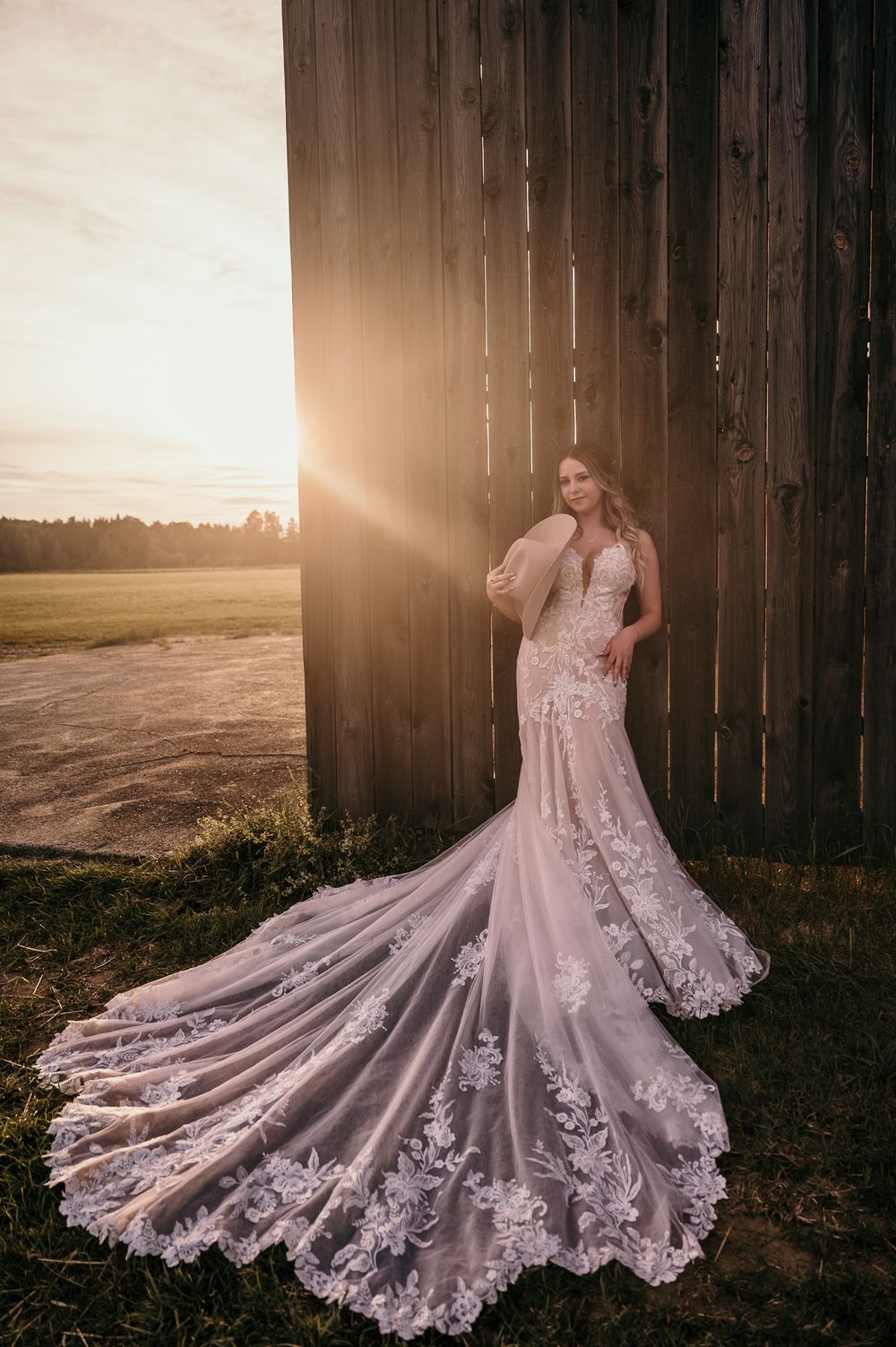 A delicate lace wedding dress is beautifully displayed against a wooden fence at sunset with a dramatic flowing train.
