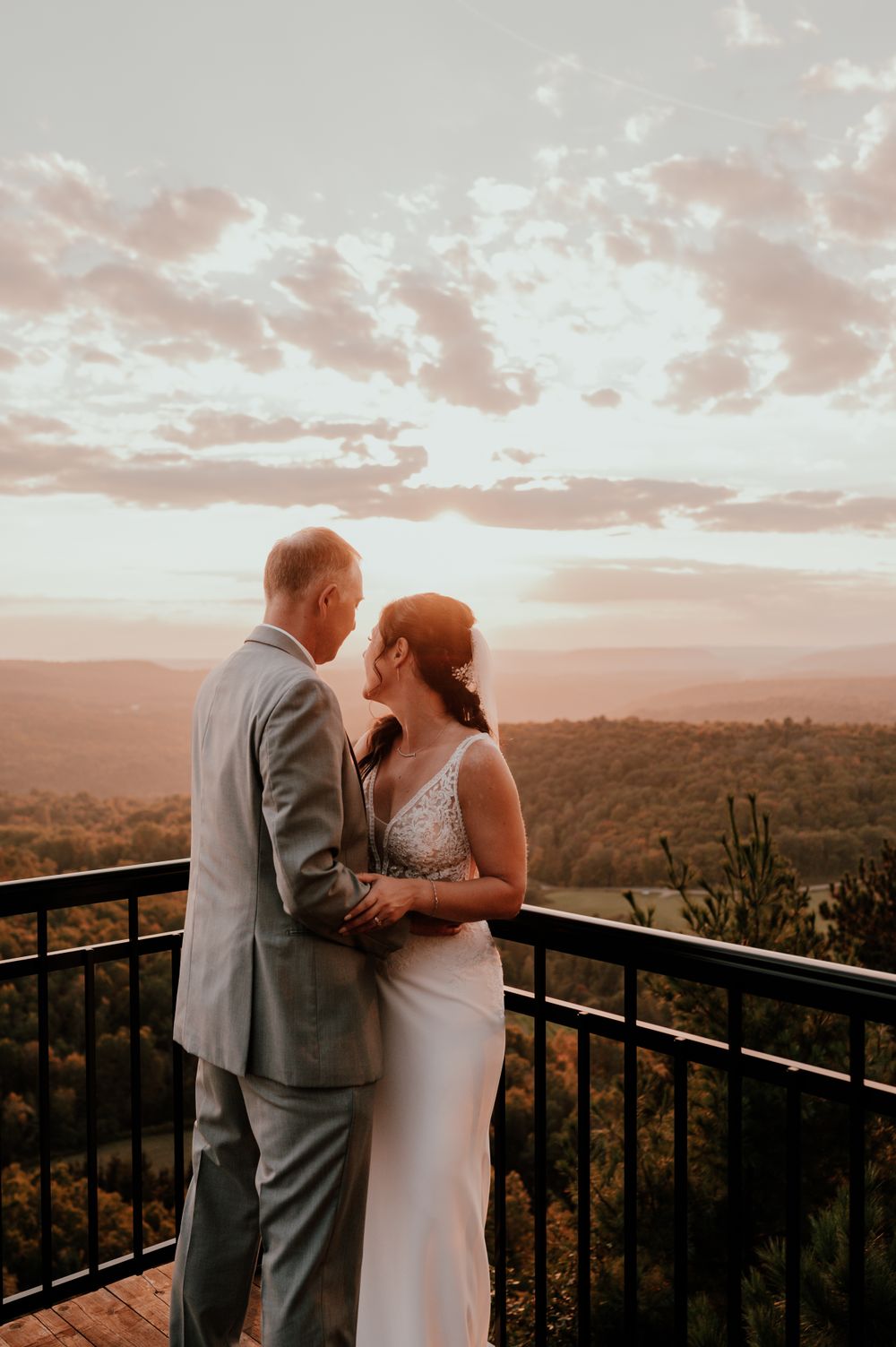 A romantic wedding couple embraces on a balcony overlooking a scenic mountain landscape at sunset.