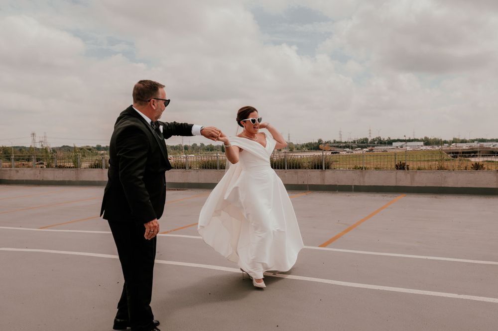 A couple in wedding attire dances joyfully on an empty rooftop parking lot under cloudy skies.