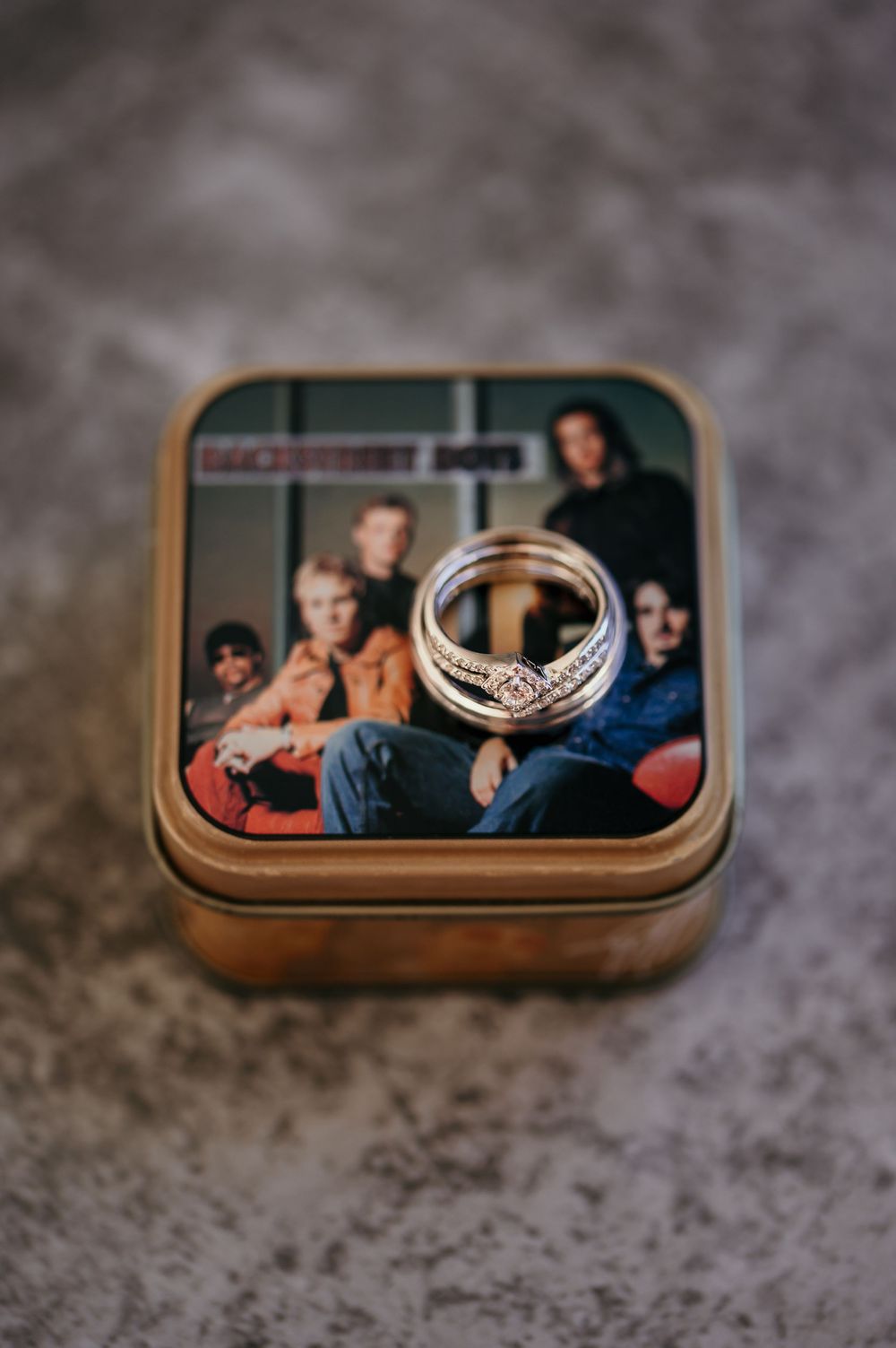 Wedding rings rest on a small brown box containing a photograph on a textured gray surface.