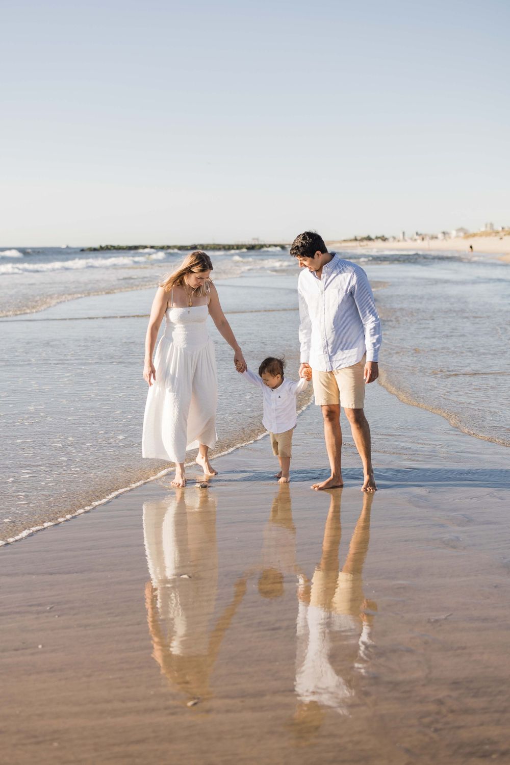 Family in white and beige clothing walking together and drawing in wet sand on a sunny beach at sunset.