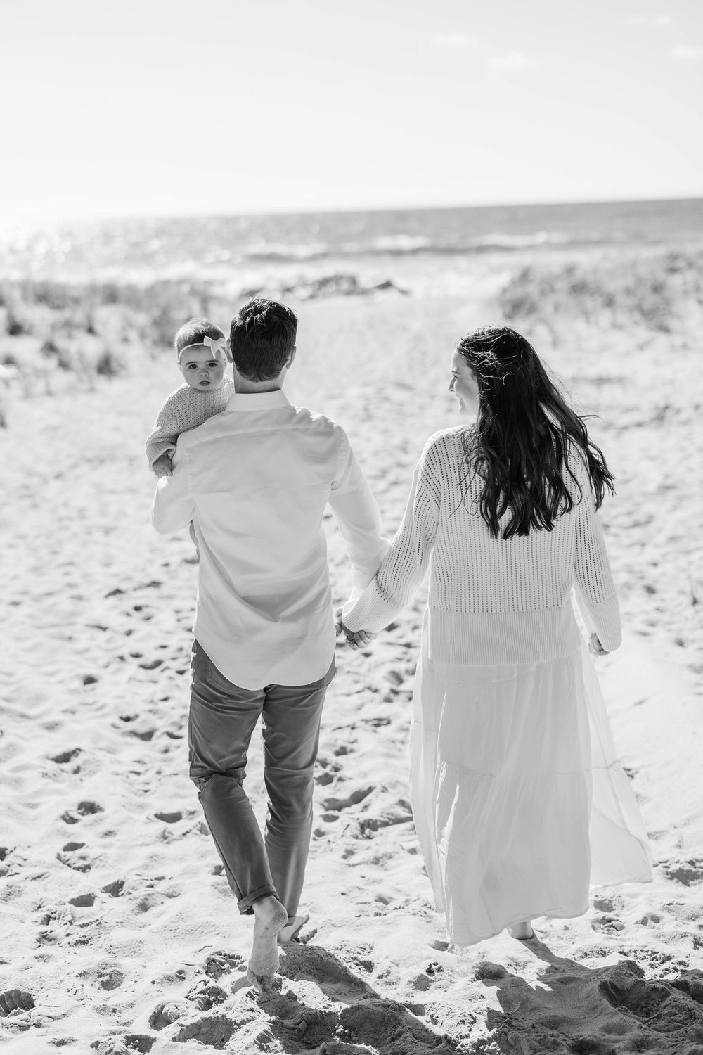 A couple in white outfits walking hand in hand on a sandy beach in black and white photography.