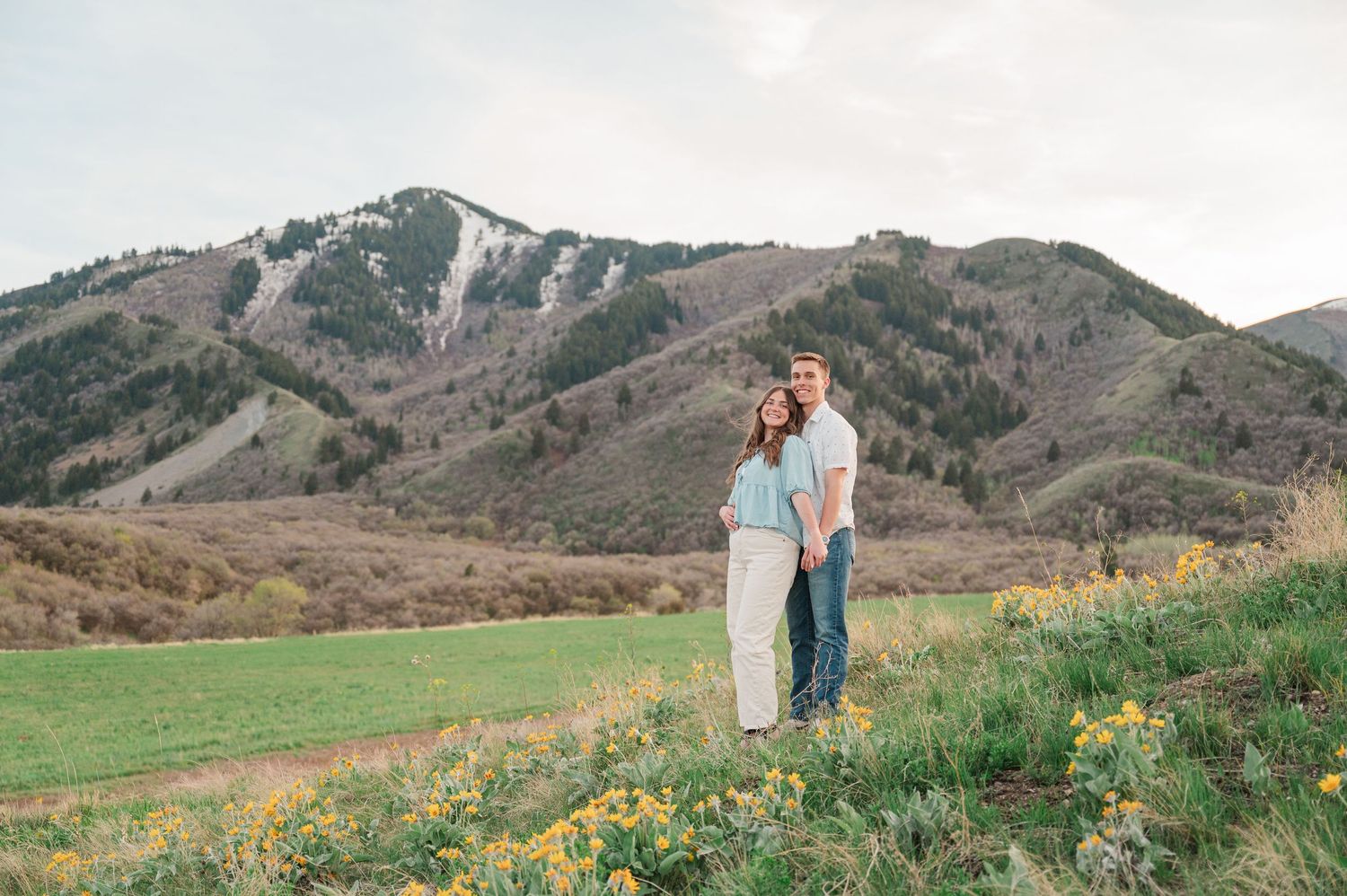 Couple embracing in a green meadow with wildflowers and a scenic mountain backdrop under a soft cloudy sky.