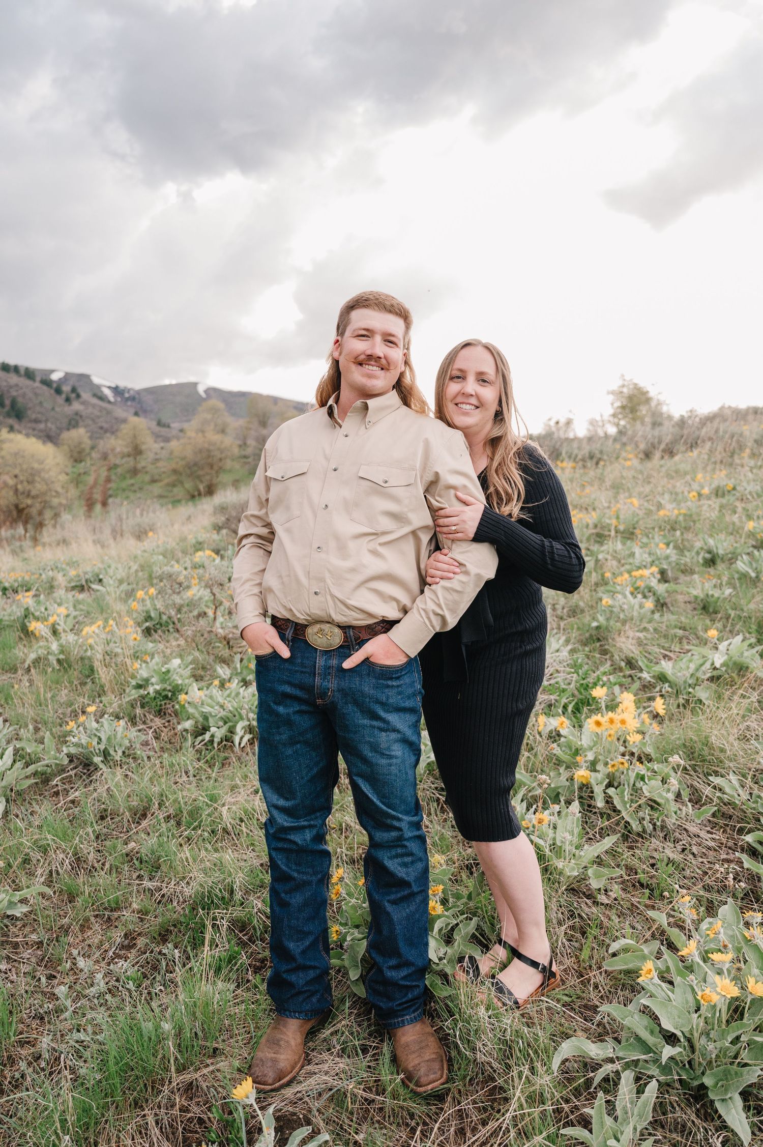 A couple stands together in a field of wildflowers with mountains in the background on an overcast day.