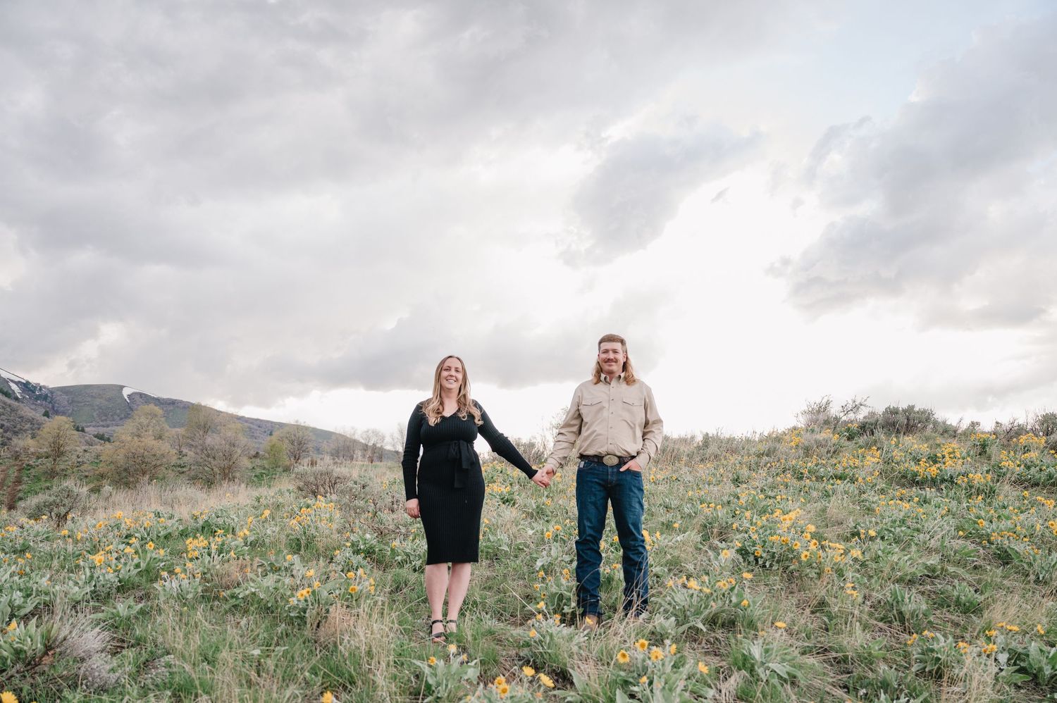 Two people hold hands while standing in a grassy mountain meadow on an overcast day.