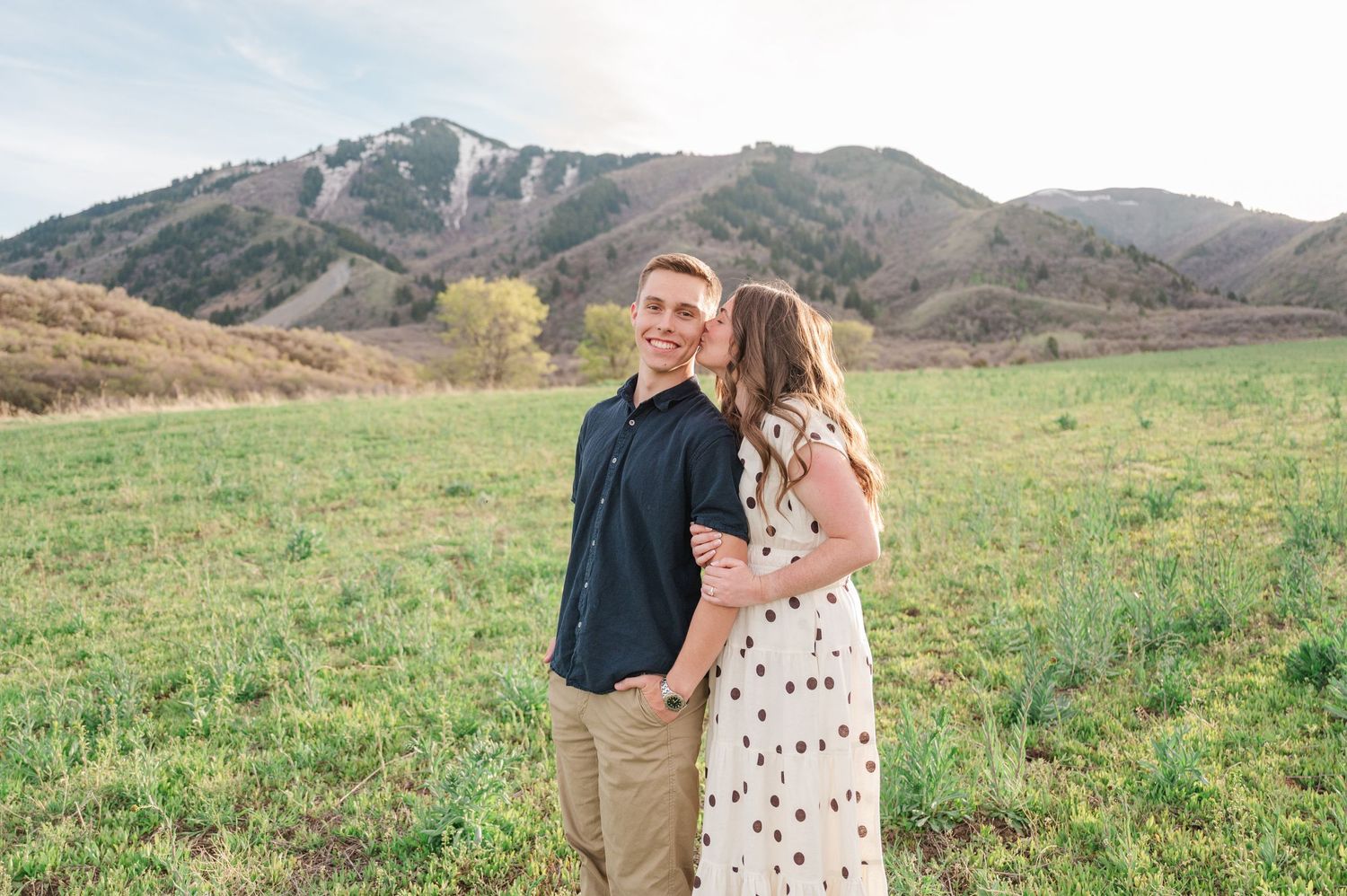 A couple shares a romantic kiss in a lush green field with scenic mountain backdrop during golden hour.