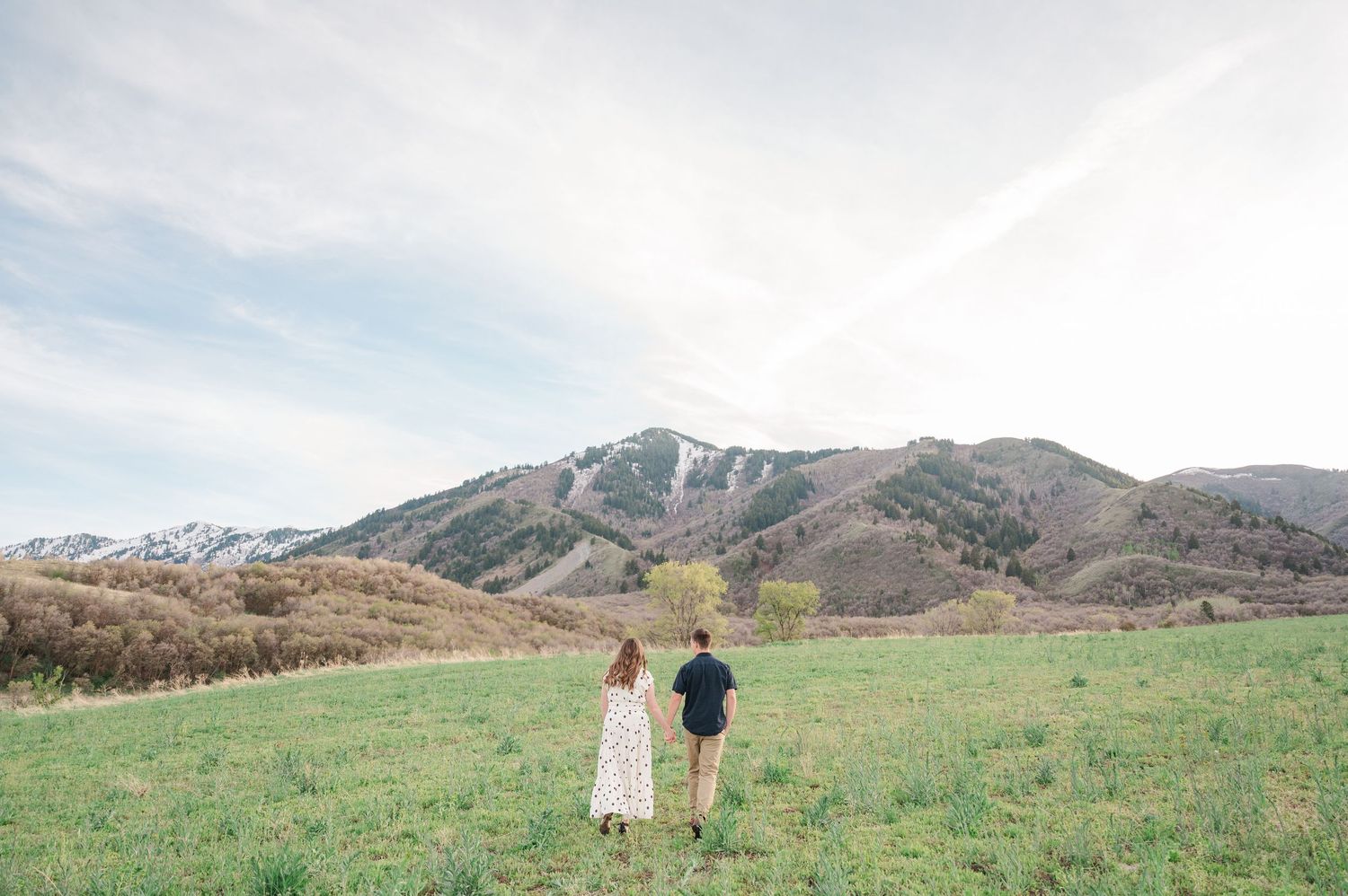 Couple holding hands walking through green meadow with snow-capped mountain backdrop under soft cloudy sky.