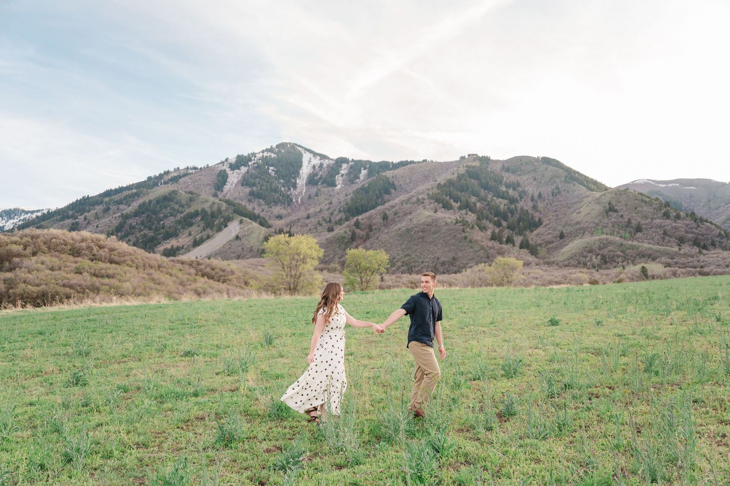 Couple holding hands walking through green meadow with mountain ski resort backdrop during spring engagement photo session.