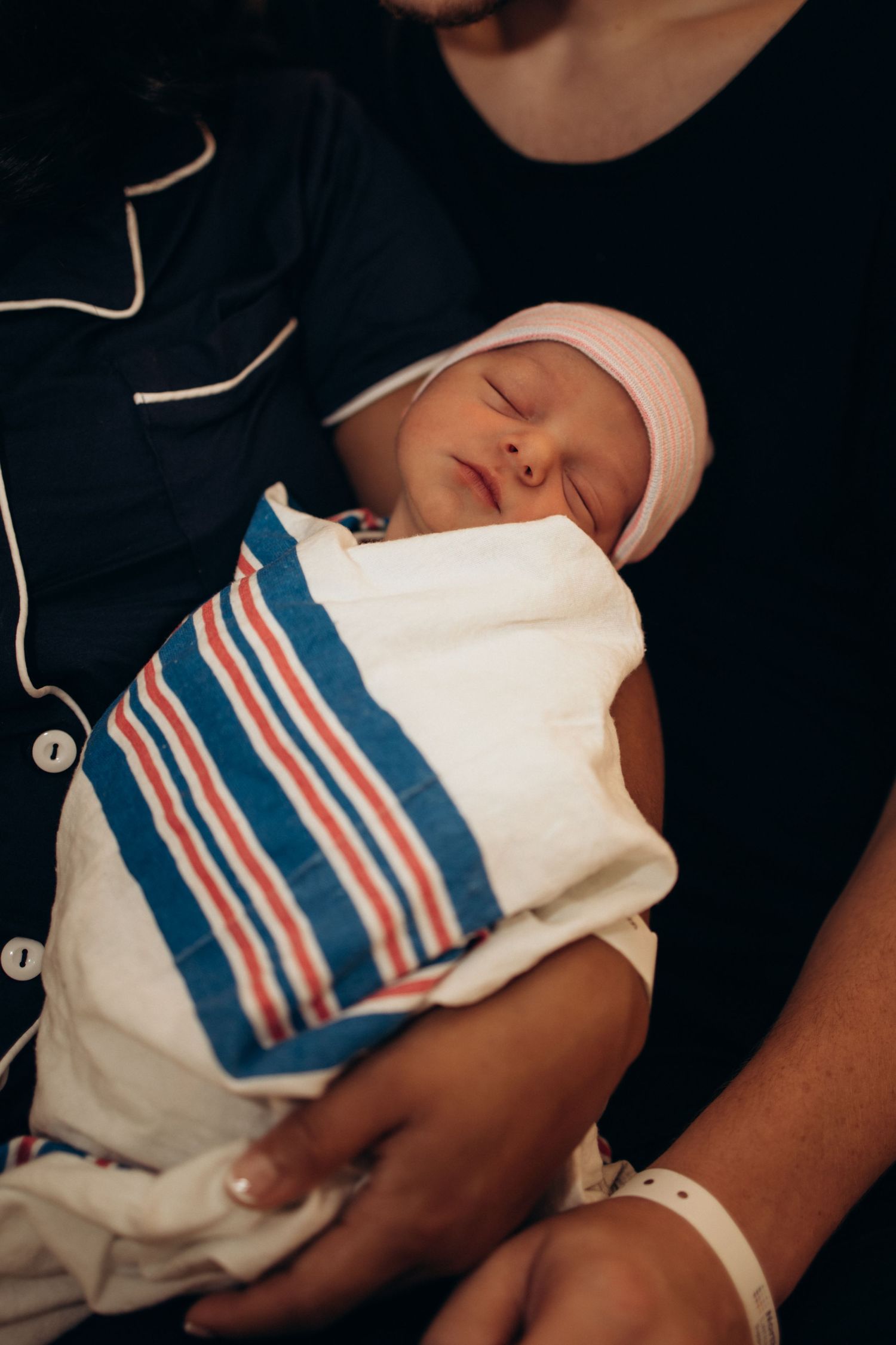 Peaceful newborn baby swaddled in striped hospital blanket with pink hat sleeping soundly.
