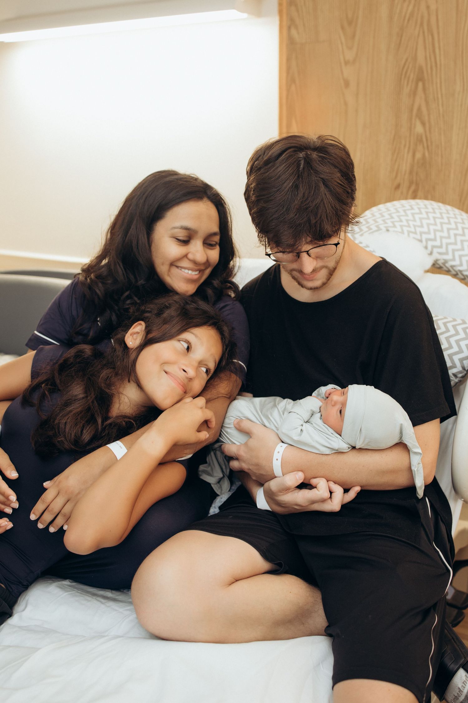 New parents and older sibling admire their newborn baby together in a hospital room.