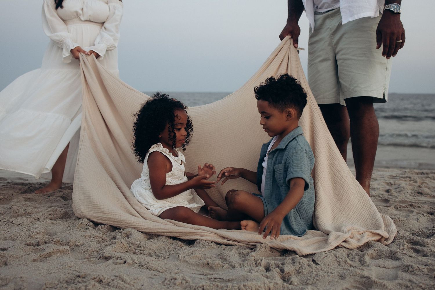 Two young children play together under a blanket tent on a sandy beach at dusk.