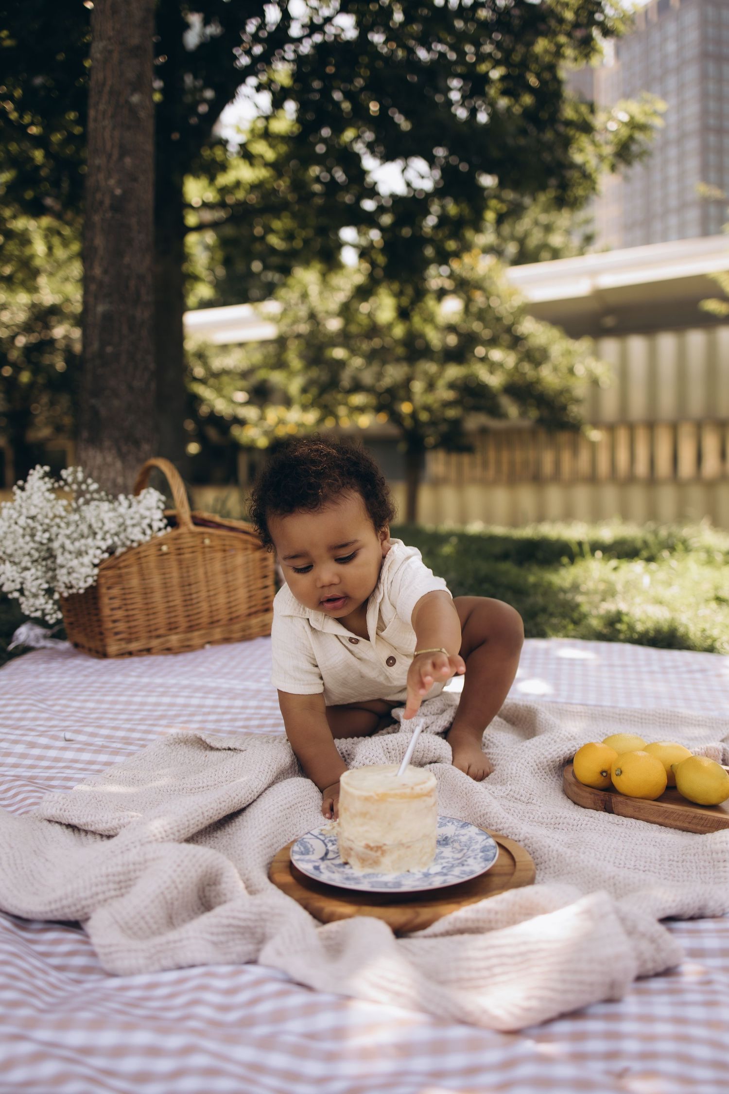 Adorable baby explores first birthday smash cake on a picnic blanket in a sunny backyard.