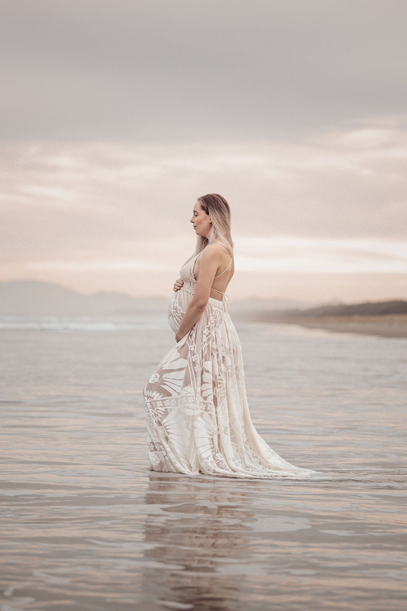 Expectant mother in flowing lace dress standing in shallow water at sunset with mountains in background.