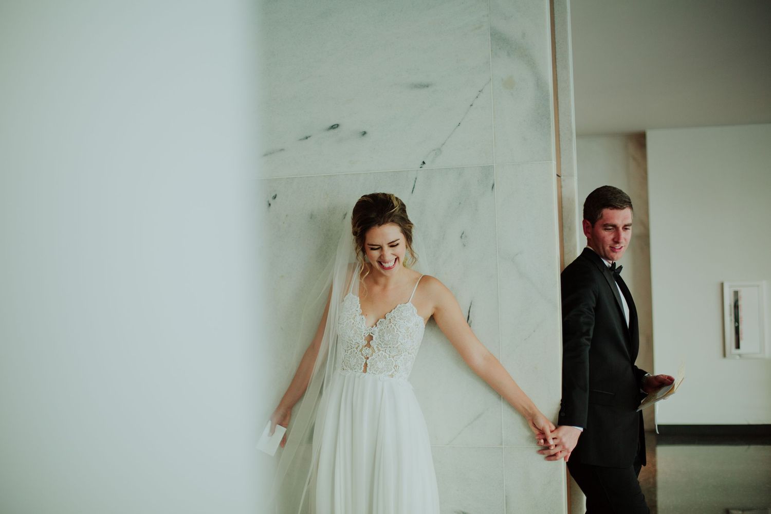 A first look wedding moment with couple separated by wall holding hands.