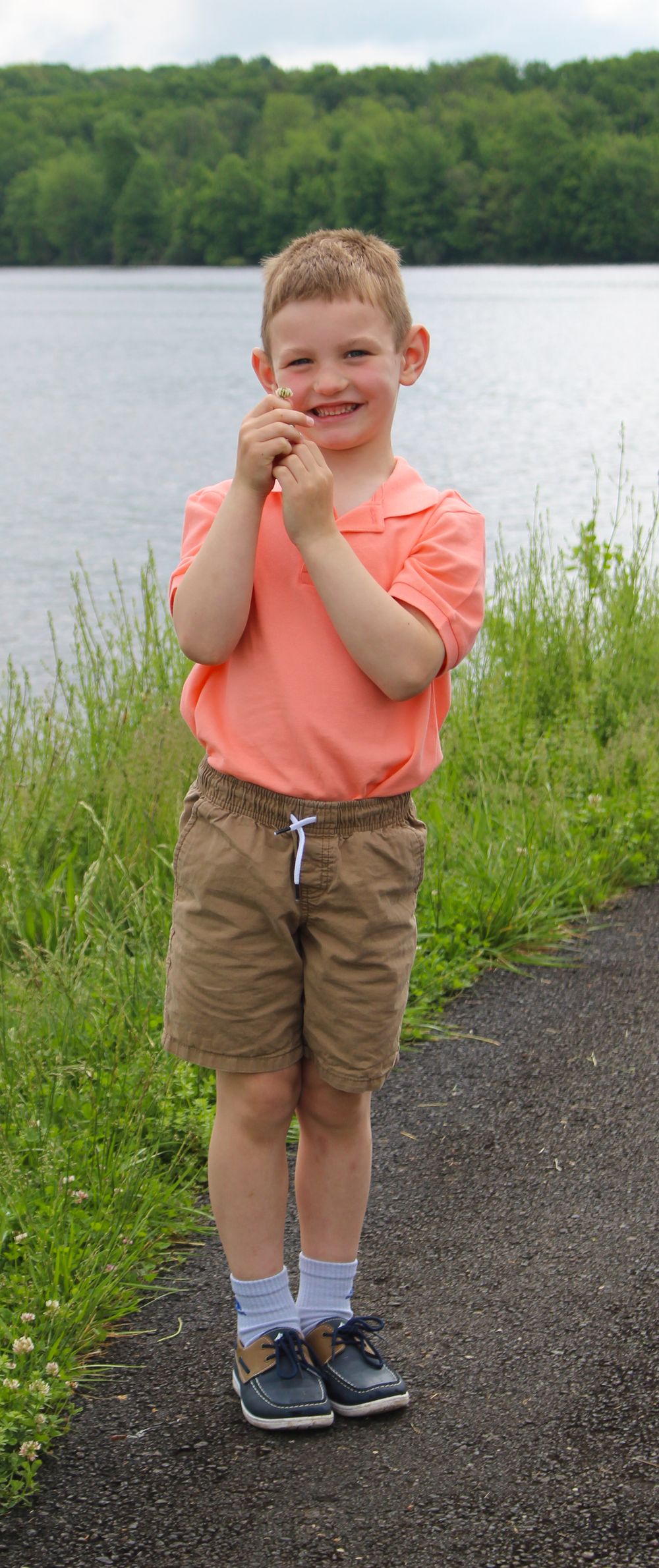 A child in coral polo and khaki shorts stands on a lakeside path with clasped hands.