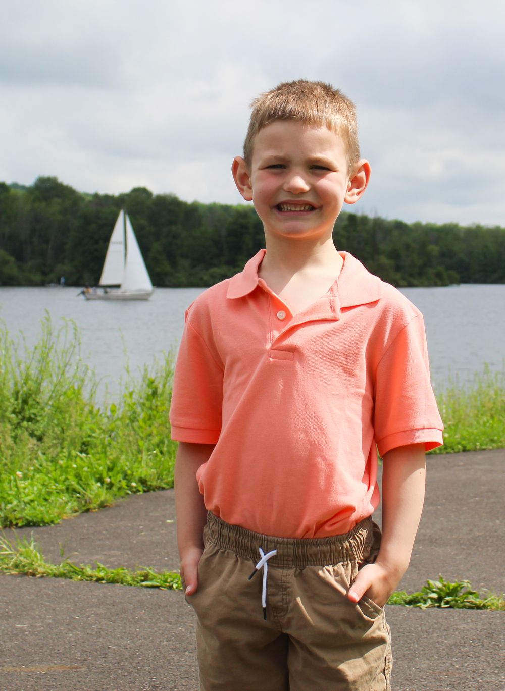 A child in a coral polo shirt smiles next to a lake with a sailboat visible in the background.