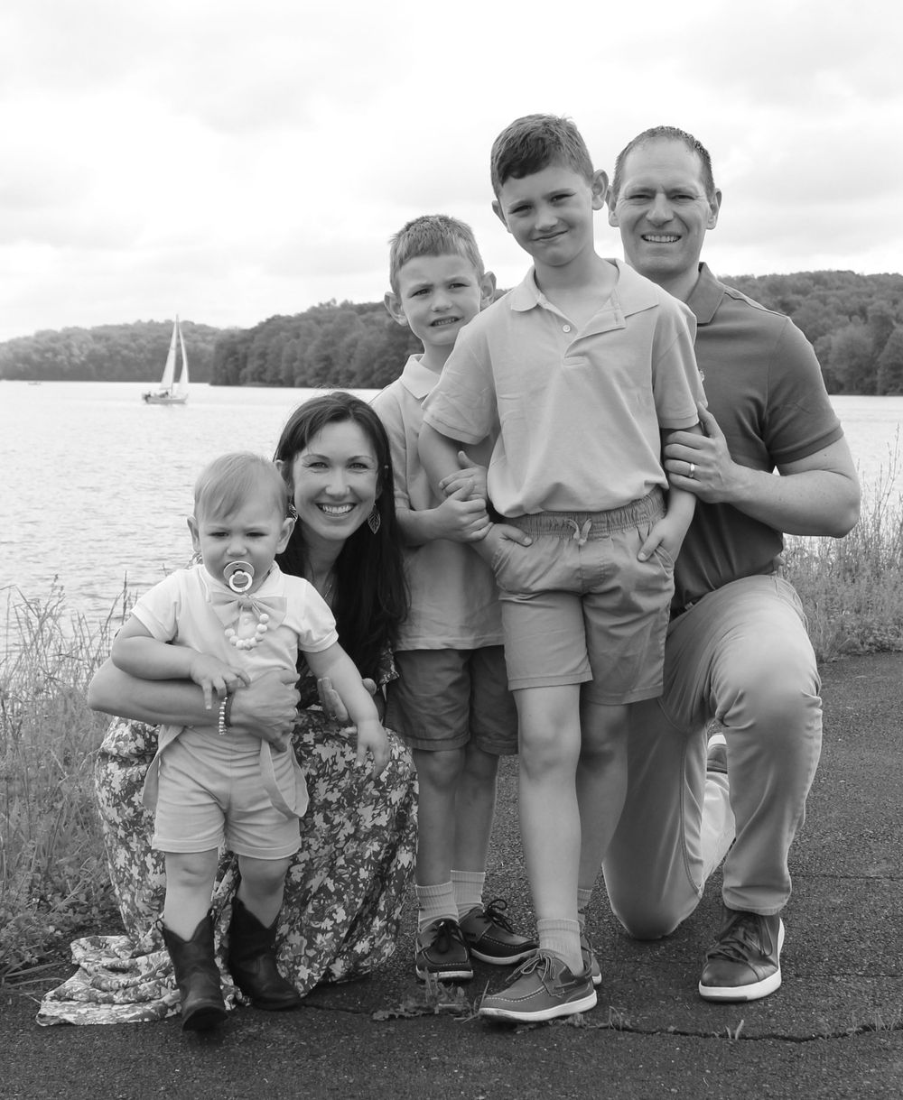 Black and white portrait of a family group posed by a lake with a sailboat in the distance.