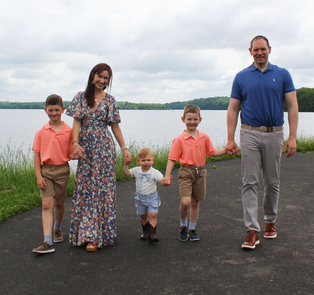 Family standing on a waterfront path with sailboat and lake scenery behind them.