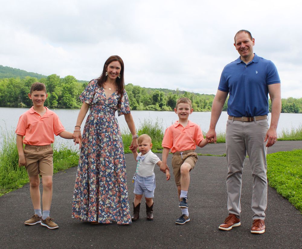 A family of five holds hands while walking along a lakeside path on a cloudy day.