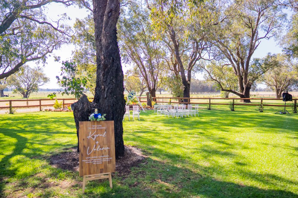 Outdoor wedding ceremony setup with wooden signage and white chairs under large trees.