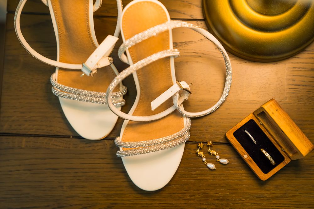 White strappy wedding sandals and ring box displayed on dark wooden surface.