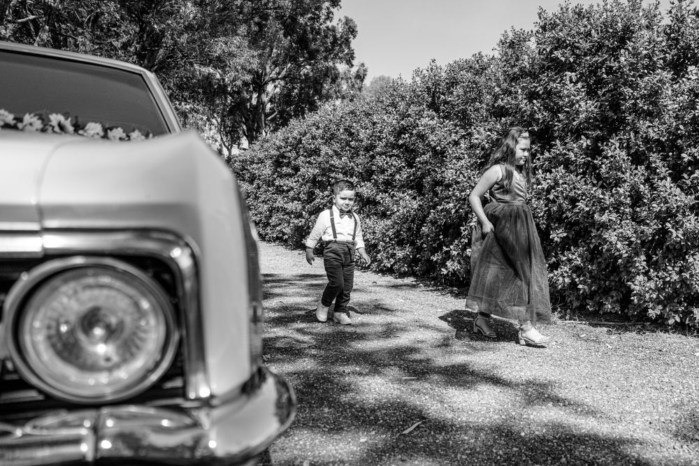 A vintage car sits in foreground of a countryside path with wedding party in background.