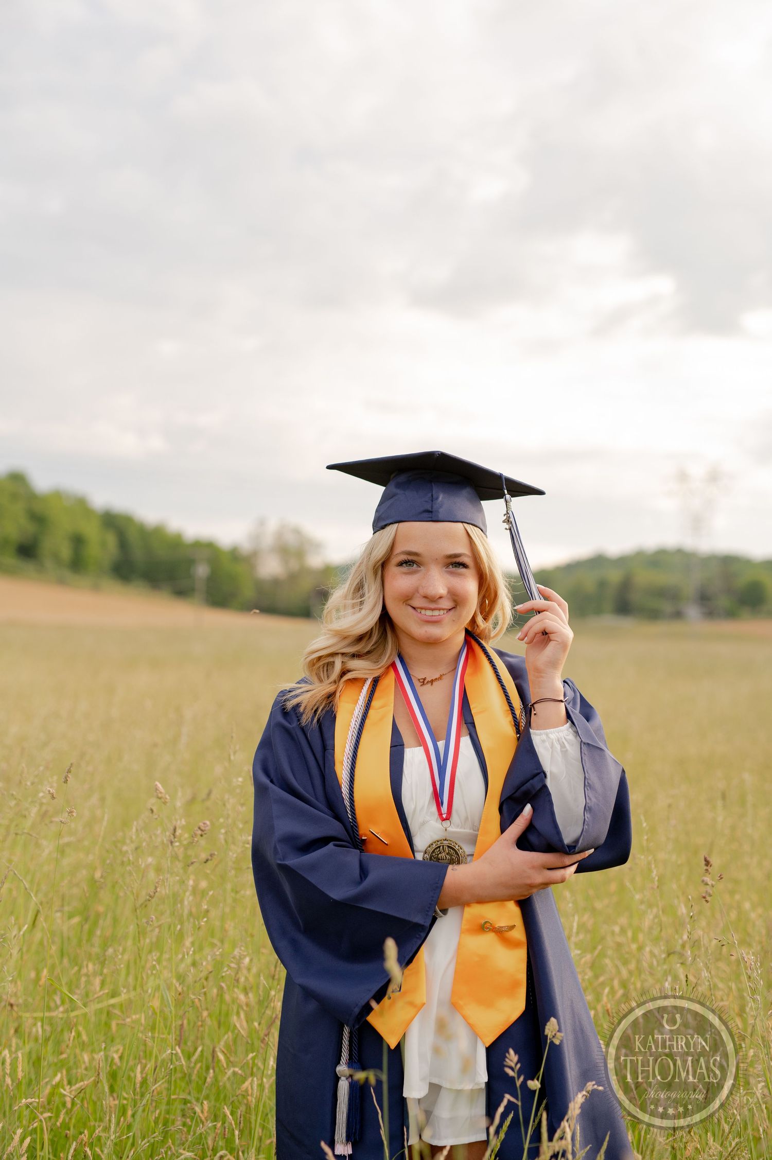 Graduate wearing blue cap and gown with gold stole and medal poses in sunny field.