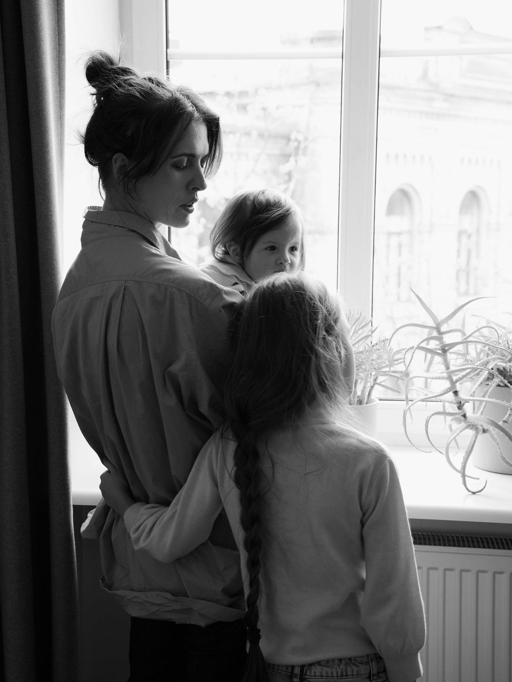 Mother and two daughters are joyfully standing next to the window.