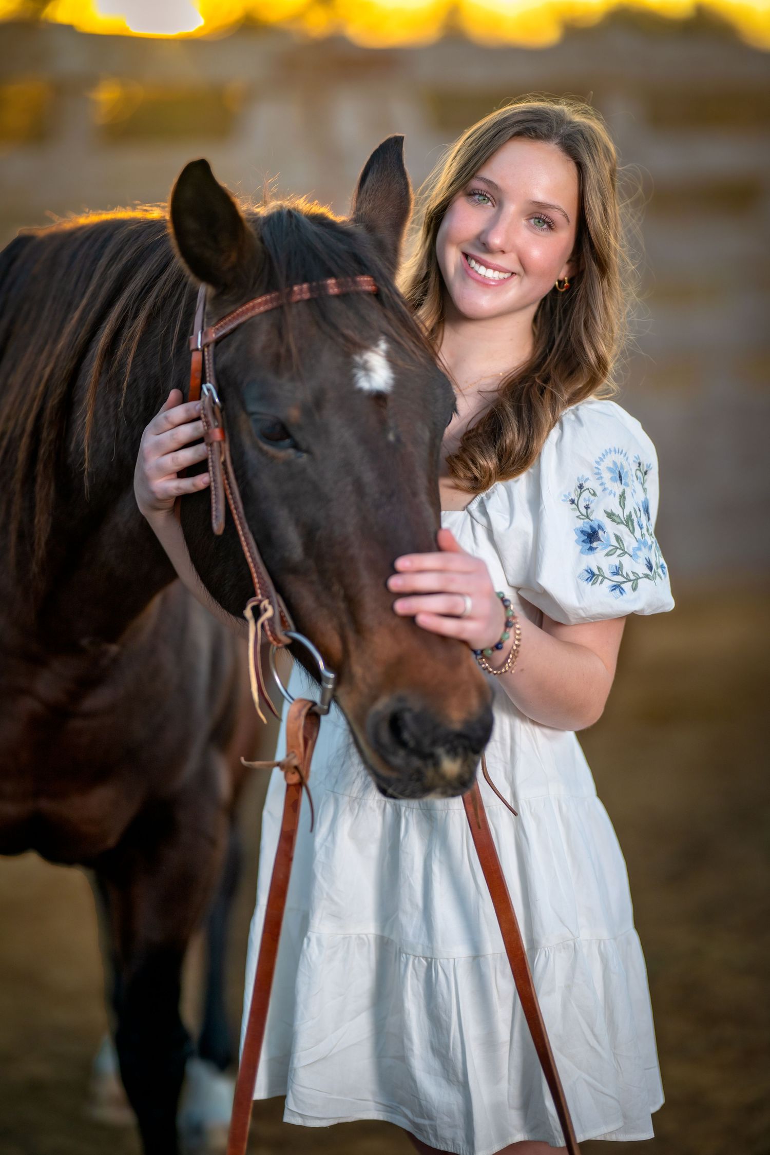 A beautiful dark brown horse poses with someone wearing a white dress during golden hour sunset lighting.