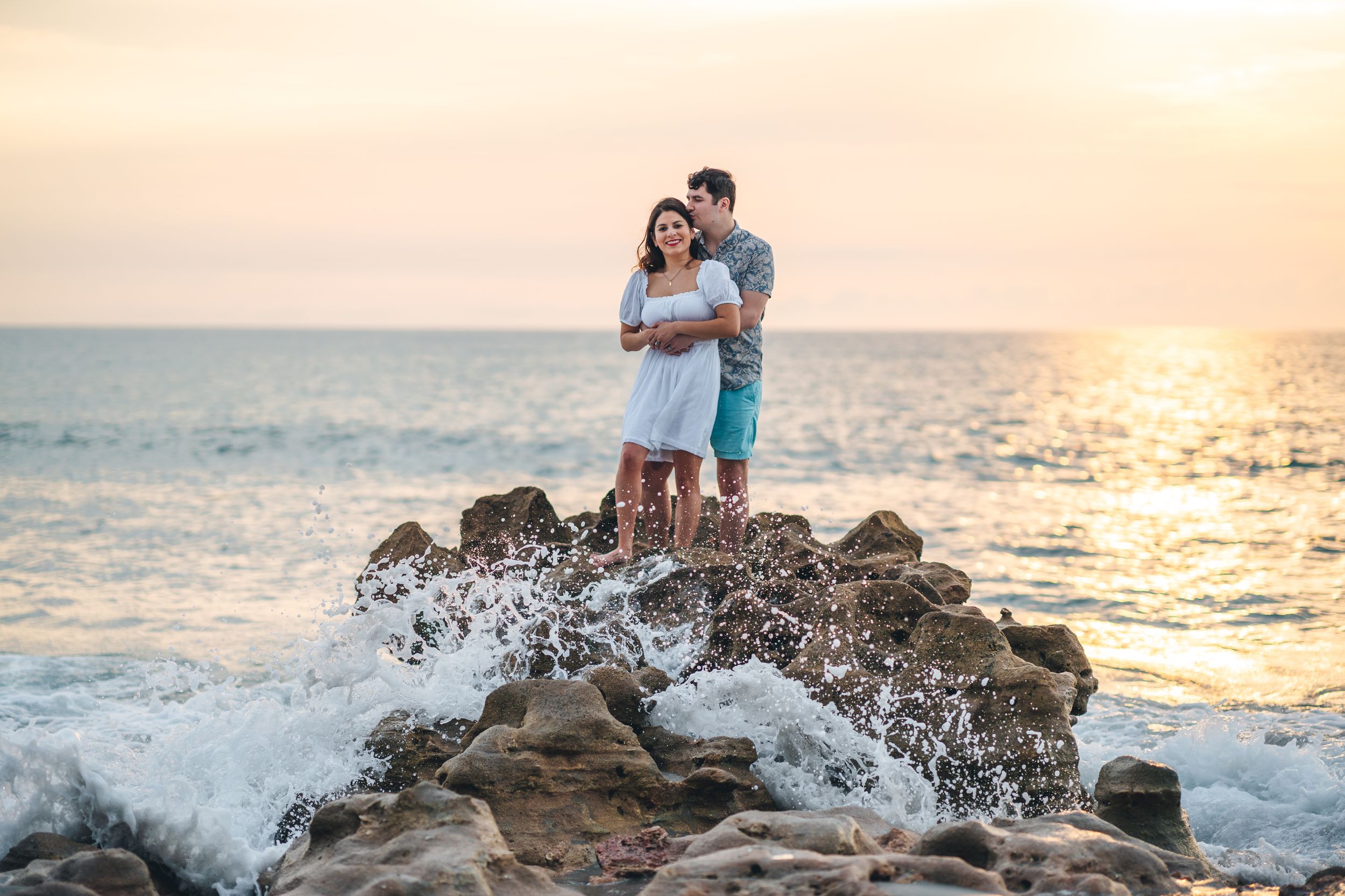 Romantic couple embracing on rocky beach at sunset with ocean waves crashing.