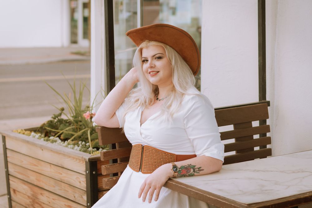 Blonde woman smiling while adjusting hat for college senior photos at Milk Bottle on Garland bench - Gabbie Rose Photo