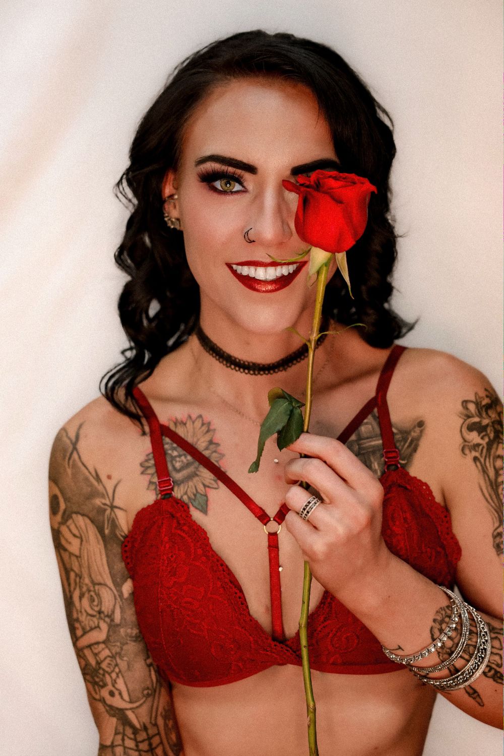 Glamour portrait with red rose featuring tattoos, red lingerie, and silver bracelets against light background.