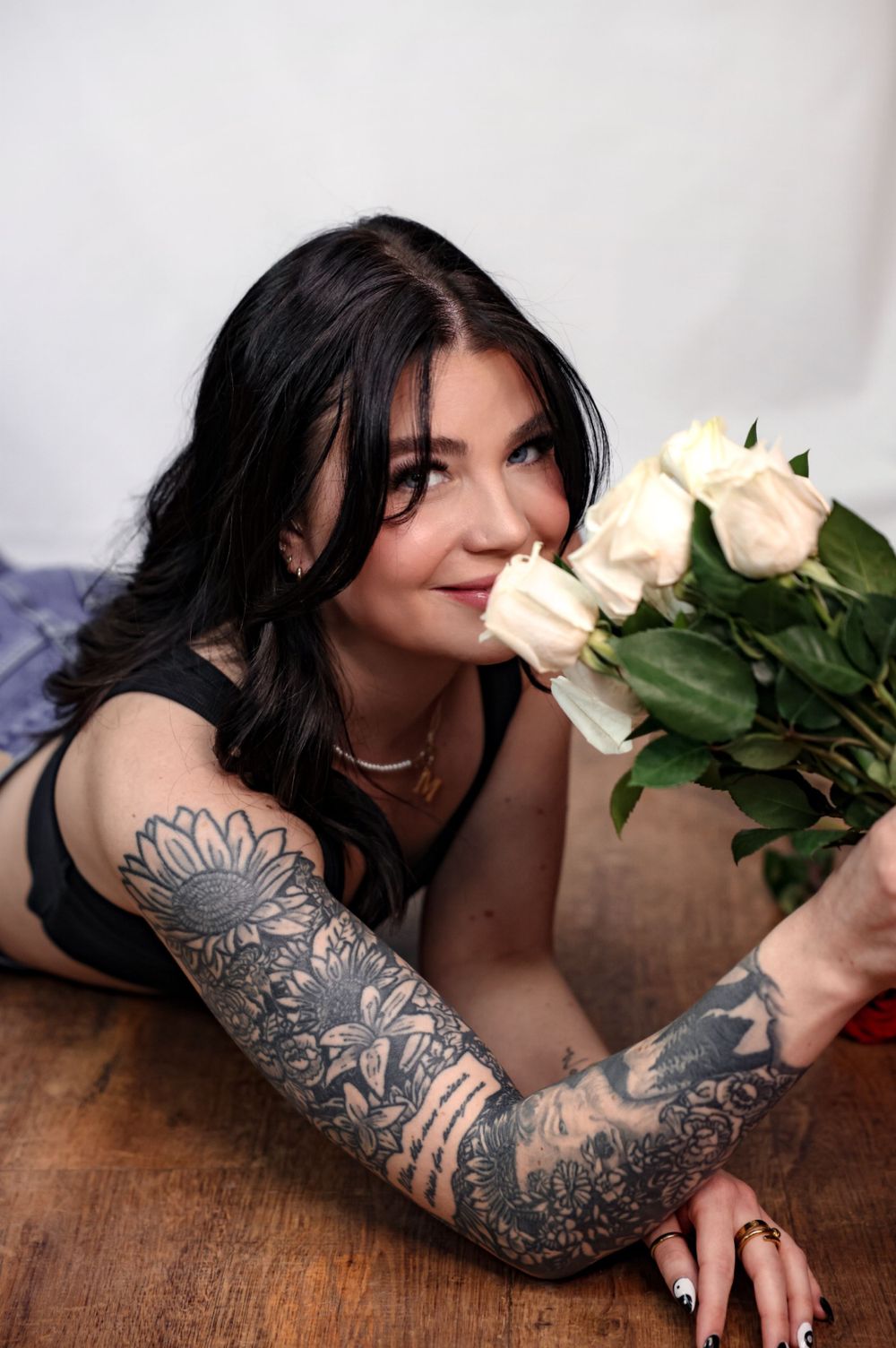 Moody portrait with white roses and ornate floral sleeve tattoo on wooden table.