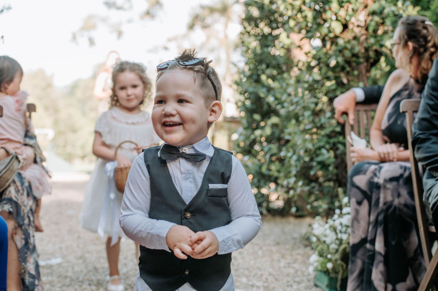 Young ring bearer in vest and bow tie smiling during outdoor wedding celebration.
