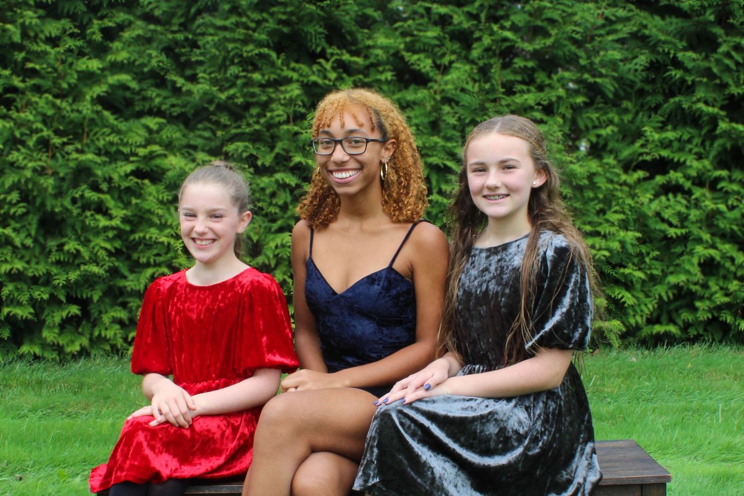 Three people in formal attire seated on a bench with greenery in background.