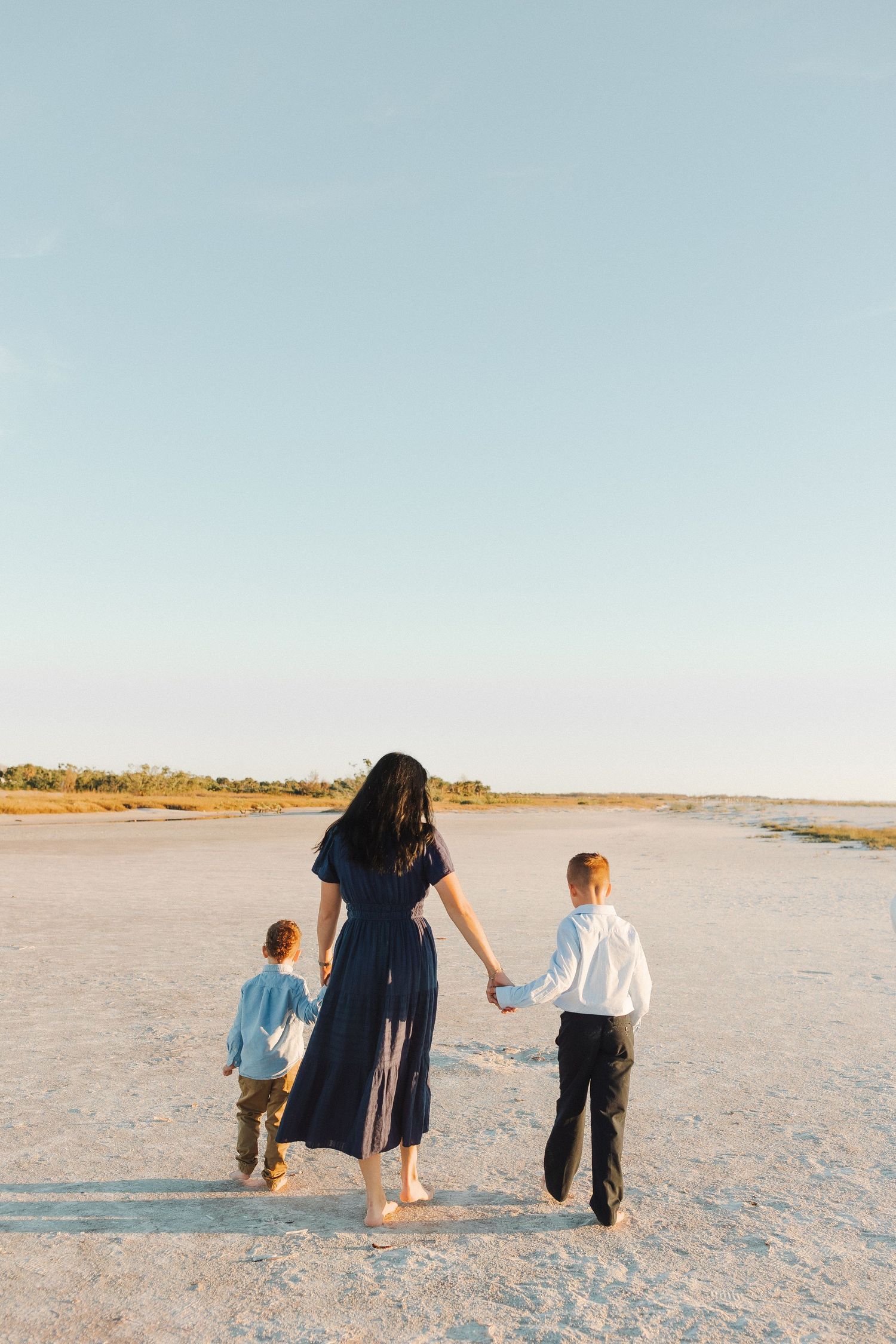 A mother in a blue dress walks hand-in-hand with two children across a sandy beach at sunset.